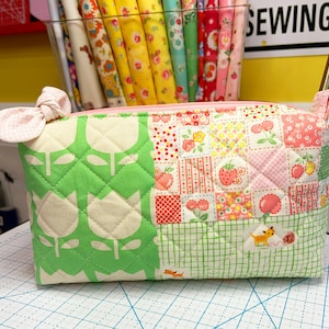 May include: A quilted fabric pouch with a pink zipper and a bow. The pouch features a green tulip pattern, patchwork squares with strawberries and flowers, and a green gingham bottom. The word "SEWING" is visible in the background.