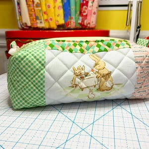May include: A quilted fabric pencil case with a green and white gingham pattern on one end and a white diamond pattern on the other. The center features a bunny illustration. The zipper is yellow.