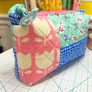 May include: Quilted fabric pouch with a pink zipper. The pouch features a patchwork design with a pink tulip pattern, floral print, and blue gingham. A fabric bow is attached to the side. The pouch is likely used for cosmetics or small items.