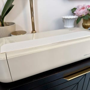 May include: Cream-colored Cricut machine with a white paper strip on top. The machine is on a black cabinet with gold handles. The Cricut logo is visible on the machine. A gold lamp and a potted plant are in the background.