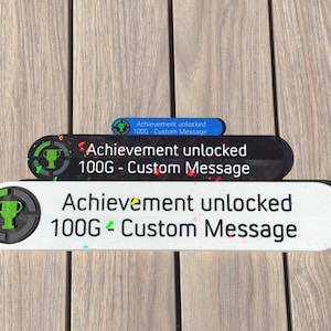 May include: Three achievement badges with rounded edges. The badges are white, black, and blue, each with the text "Achievement unlocked 100G - Custom Message." Each badge has a green trophy icon.