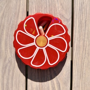May include: A circular red pill organizer with a white flower-shaped design on the lid. The lid features a rotating mechanism with a single opening that reveals a small white capsule inside. The container has a scalloped edge and a gold center.