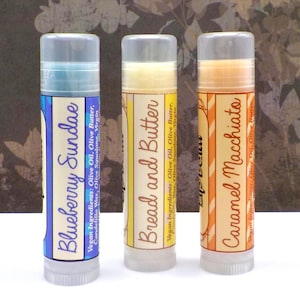 May include: Three lip balm tubes with labels reading "Blueberry Sundae", "Bread and Butter", and "Caramel Macchiato". The tubes are clear with gray caps. The labels list vegan ingredients.