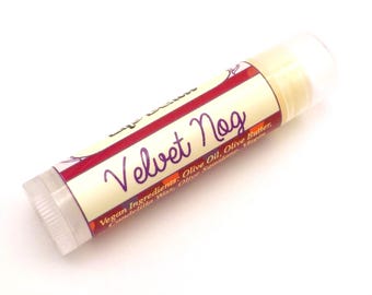 May include: A cylindrical lip balm tube with a cream-coloured label featuring the purple text Velvet Nog. The label includes red accents and lists vegan ingredients such as olive oil, olive butter, and candelilla wax in small, dark-coloured print.