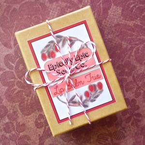 May include: A brown box tied with red and white twine. The box has a label that says "Epically Epic Soap Co." and "Lip Balm Trio".