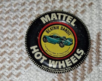 May include: A vintage Hot Wheels Beatnik Bandit button. The button features a black rim with the words "MATTEL HOT WHEELS" in white. The center has a yellow circle with a teal car graphic and the words "BEATNIK BANDIT".