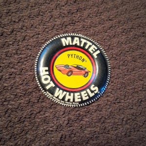 May include: A round, black Hot Wheels button featuring a red car on a yellow background. The button has the text "MATTEL" and "HOT WHEELS" in white, with "PYTHON" in yellow. The button is ringed with a white and black pattern.
