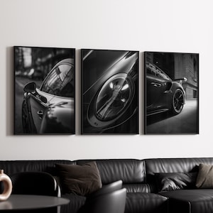 May include: Set of three framed black and white prints featuring close-up shots of a sports car. The images highlight the car's sleek design, including headlights, side mirrors, and body contours. The prints are displayed above a black leather sofa.