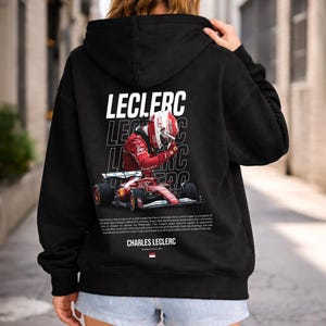 Könnte beinhalten: Schwarzer Kapuzenpullover mit einer Grafik von Charles Leclerc in rotem Rennanzug und Helm, mit dem Namen "LECLERC" in weißen Blockbuchstaben. Der Kapuzenpullover enthält auch ein Formel-1-Rennwagen und Text unten.