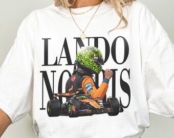 May include: White t-shirt featuring a graphic of a Formula 1 race car and driver. The text "LANDO NORRIS" is printed in large black letters. The driver is wearing a green and black helmet and orange racing suit.