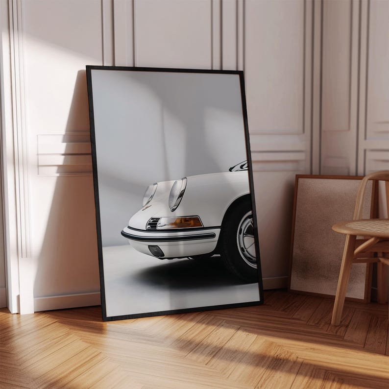 May include: A white classic car with a black bumper and black wheels. The car is photographed from the front, showing the headlights and the front grill. The car is in a room with white walls and wooden floors.