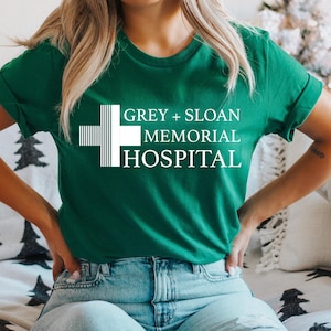 May include: A woman wearing a green t-shirt with a white graphic of the Grey Sloan Memorial Hospital logo. The logo features a stylized white cross with the text "GREY + SLOAN MEMORIAL HOSPITAL".