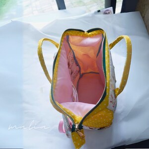 May include: Open view of a handmade backpack with a yellow polka dot exterior and a pink interior. The bag has a zippered closure, yellow handles, and a small pink strap. The interior features multiple compartments and a floral pattern.