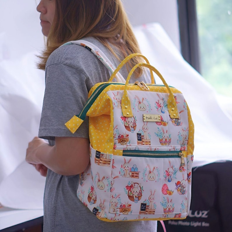 May include: A white backpack with a repeating pattern of bunnies, Easter eggs, and floral accents. The bag has yellow polka dot and solid yellow trim, green zippers, and gold hardware. The bag has a top handle and adjustable straps.