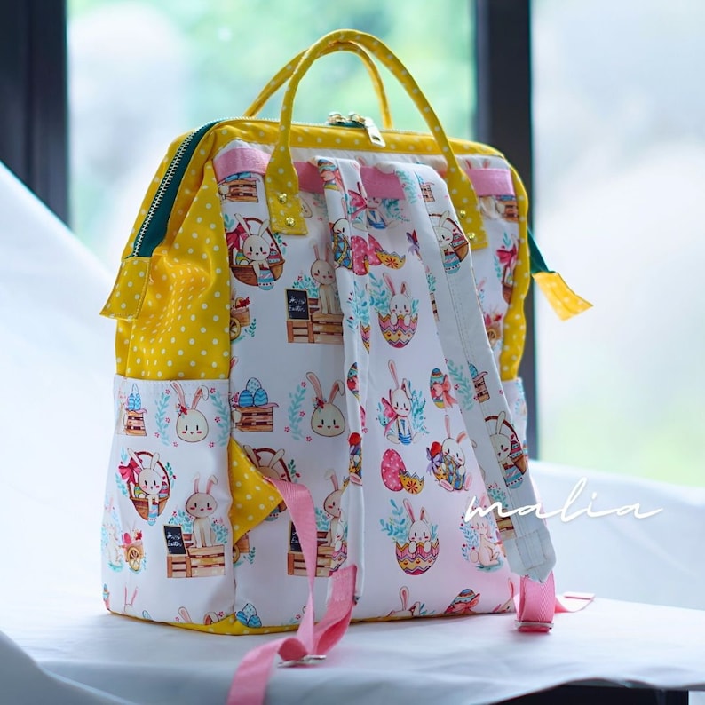 May include: A square backpack with a yellow polka dot pattern and a white Easter-themed print featuring bunnies, eggs, and baskets. The bag has pink straps, a yellow handle, and a green zipper.