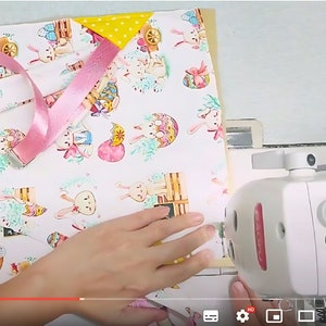 May include: A sewing machine stitching fabric with an Easter bunny print. The fabric features bunnies, Easter eggs, and floral accents on a white background. A pink ribbon with a silver buckle is laid across the fabric. A yellow polka dot fabric is also visible.