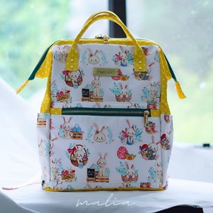 May include: White backpack with an Easter-themed pattern featuring bunnies, eggs, and baskets. The bag has yellow polka dot accents, a top handle, and a front zipper pocket. The brand name "malia" is visible at the bottom.