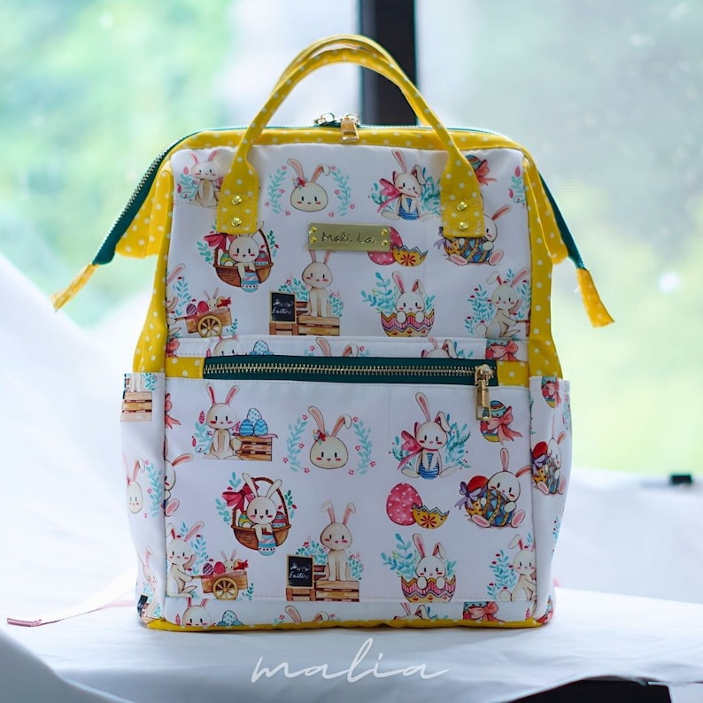 May include: White backpack with an Easter-themed pattern featuring bunnies, eggs, and baskets. The bag has yellow polka dot accents, a top handle, and a front zipper pocket. The brand name "malia" is visible at the bottom.