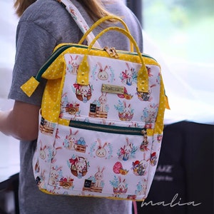 May include: A white and yellow backpack with a repeating pattern of Easter bunnies, eggs, and floral accents. The backpack features yellow polka dot accents, a gold zipper, and a small gold nameplate that reads "Mali. La."