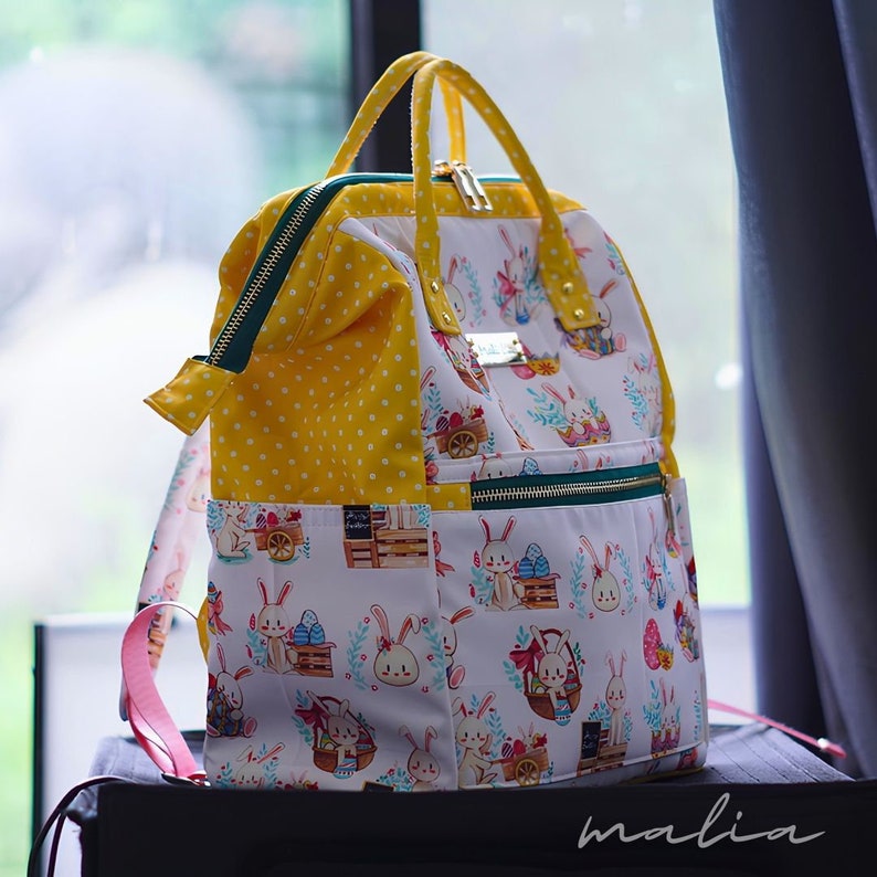 May include: A yellow and white backpack with a whimsical Easter bunny print. The bag features a yellow polka dot pattern, green zippers, and a pink strap. The bag has a top handle and a front pocket.