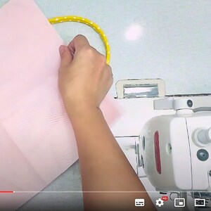 May include: A close-up shot of a sewing project in progress. A hand holds a piece of light pink fabric near a yellow polka-dotted handle. A white sewing machine is in the background, suggesting a crafting or DIY theme.
