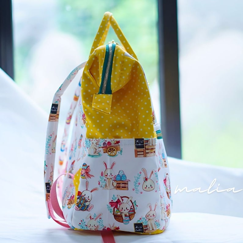 May include: A white backpack with a colorful Easter bunny print. The bag has a yellow polka dot interior and a green zipper. The straps are white and pink. The bag has a pocket with a bunny and Easter egg design.