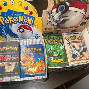 Puede incluir: Una exhibición de paquetes de cartas coleccionables Pokémon vintage, incluidas las ediciones Team Rocket y Jungle Booster. Las cajas presentan el logotipo de Pokémon y el texto "1st Edition Limited Printing". Los paquetes están sellados y presentan varios personajes Pokémon.