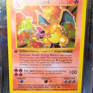 May include: A Charizard Pokémon card with a yellow border. The card features an illustration of a fiery orange dragon with outstretched wings. Text includes "Charizard", "120 HP", and "Fire Spin". The card is in a protective case.