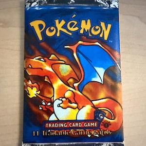May include: Sealed Pokémon Trading Card Game pack. The blue pack features the Pokémon logo and a Charizard illustration in orange and yellow. Text reads "Trading Card Game" and "11 Tradable Game Cards."