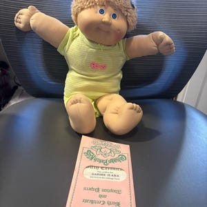 May include: A Cabbage Patch Kids doll with curly blonde hair and blue eyes, wearing a light yellow striped onesie with a pink cherry graphic. Included is a pink envelope labeled Cabbage Patch Kids Adoption Papers and Birth Certificate.