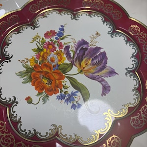 Puede incluir: Una bandeja decorativa de metal con un diseño floral. La bandeja presenta un interior blanco con un ramo de flores naranjas, moradas y azules. El borde es burdeos con detalles de filigrana dorada. La bandeja tiene un borde festoneado.