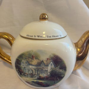 May include: White ceramic teapot with gold-colored handle, spout, and lid accents. The teapot features a painted scene of a house and the text "HOME IS WHERE THE HEART IS". The lid has a gold finial.