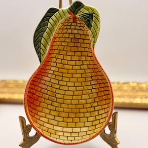 May include: A pear-shaped ceramic dish featuring a yellow and orange brick-patterned surface with green leaves at the top. The dish is displayed on a decorative gold-toned metal stand with ornate, curved legs and a vertical support.
