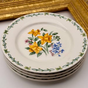 May include: A stack of four white ceramic plates. Each plate features a floral design with yellow daffodils, blue and pink flowers, and green leaves. A green vine border encircles the rim of each plate.