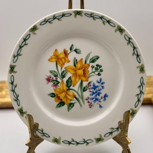 May include: A white ceramic plate with a floral design featuring yellow daffodils, blue and pink flowers, and green leaves. The plate has a green vine border and is displayed on a gold-colored stand. A decorative piece for the home.