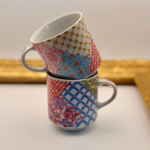 May include: Two stacked ceramic mugs with gray handles. The mugs feature a patchwork design with patterns in red, blue, pink, green, and yellow. The mugs are on a white surface with a gold-colored frame in the background.