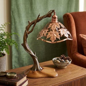 May include: A table lamp featuring a base and arm sculpted to resemble a natural tree branch with a smooth wooden foundation. The copper-toned metal lampshade is intricately cut with a repeating pattern of maple leaves throughout its surface.