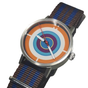 May include: A silver wristwatch with a brown and blue striped nylon band. The watch face has a white background with a concentric circle design in orange, blue, purple, and yellow.