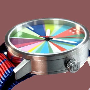 Peut inclure: Gros plan d'une montre-bracelet avec un cadran multicolore segmenté. La montre a un boîtier argenté et un bracelet à rayures rouges, blanches et bleues. Le cadran présente un arc-en-ciel de couleurs, avec des aiguilles blanches et une trotteuse rouge.