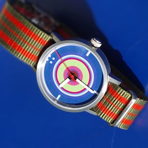 Peut inclure: Une montre-bracelet argentée avec un cadran au design de cible coloré. La montre a un bracelet en nylon rayé vert et orange.