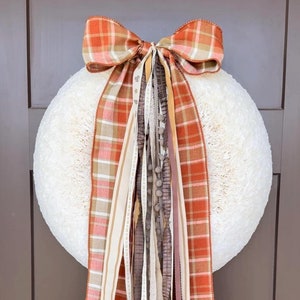 May include: A white, fluffy wreath with a large plaid bow in shades of orange, brown, and cream. The bow is tied with multiple ribbons in various textures and colours, including brown, cream, and pink.