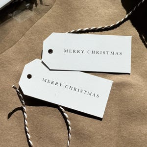 Puede incluir: Etiquetas de regalo rectangulares blancas con el texto "MERRY CHRISTMAS" impreso en negro. Las etiquetas están unidas a un rollo de cuerda marrón y blanca. Algunas etiquetas están empaquetadas en una bolsa de plástico transparente.
