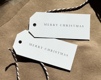 Könnte beinhalten: Weiße, rechteckige Geschenkanhänger mit dem Text "MERRY CHRISTMAS" in Schwarz gedruckt. Die Anhänger sind an einer Rolle aus braun-weißem Garn befestigt. Einige Anhänger sind in einer transparenten Plastiktüte verpackt.