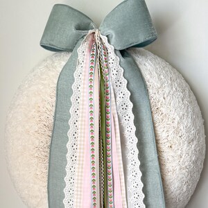 Puede incluir: Una corona redonda de color crema con un gran lazo azul polvoriento en la parte superior. Varias cintas en rosa, verde y blanco con detalles florales y de encaje cuelgan del lazo. Un artículo decorativo.
