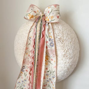 Puede incluir: Una corona redonda y blanca con un gran lazo y cintas que fluyen. El lazo y las cintas presentan un estampado floral con flores rosas, amarillas y azules. Las cintas incluyen encaje y terciopelo en tonos rosa y crema.