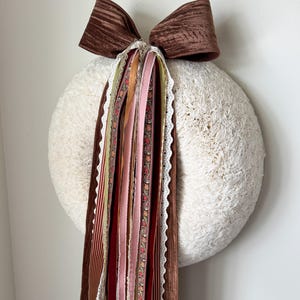 May include: A large, round, textured cream-coloured wreath adorned with a prominent brown velvet bow at the top. Cascading from the centre of the bow are multiple long, vertical ribbons in various shades of brown, pink, red, and cream, featuring diverse textures, patterns, and lace trim.