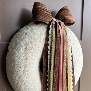 May include: A circular wreath covered in cream-coloured textured fabric, featuring a large brown velvet bow at the top. A collection of long, hanging ribbons in shades of brown, green, pink, and yellow, including lace and floral patterns, drapes down.