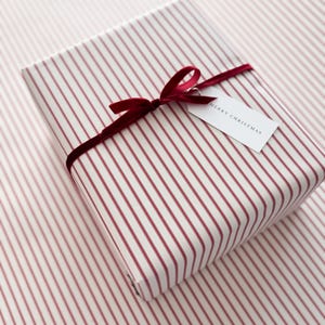 May include: A rectangular gift box wrapped in white paper with thin vertical red stripes. The package is tied with a dark red velvet ribbon bow and features a small white rectangular gift tag with the text MERRY CHRISTMAS printed on it.