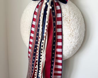 May include: A white textured wreath with a large red and white gingham bow. Red, white, and blue ribbons with stars and pom-poms hang down. A patriotic home decoration.