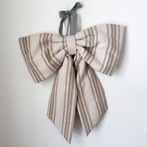 May include: A large decorative bow made of beige fabric with vertical brown stripes. The bow is suspended by a gray ribbon, creating a rustic and elegant aesthetic. The bow is a decorative accent for home decor.